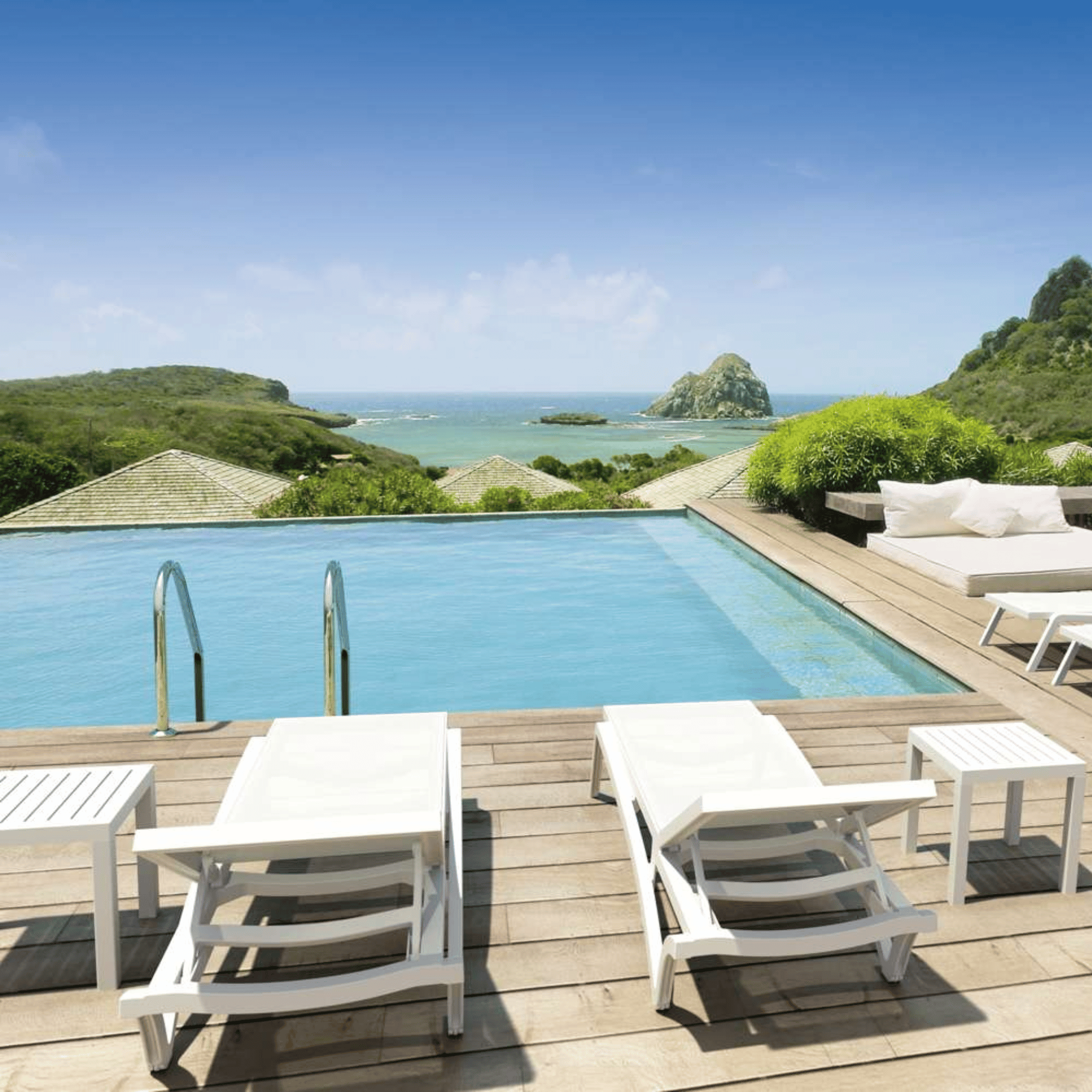 A luxury outdoor infinity pool overlooking a tropical ocean and rocky island landscape, with white lounge chairs, tables, and a wooden deck in the foreground.