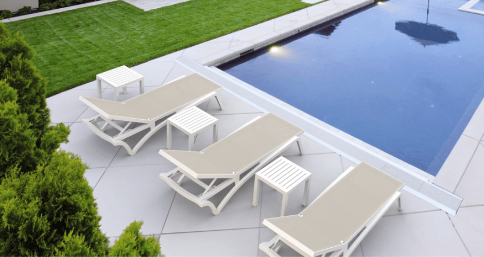 3 white resin sunlounges and side tables sitting around a blue pool
