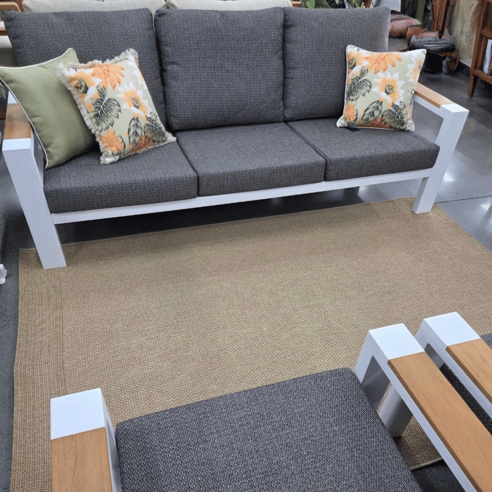 A modern outdoor Aspen three-seat sofa with dark gry textured cushions and a white frame, styled with two decorative throw pillows on a beige woven rug in a furniture showroom.