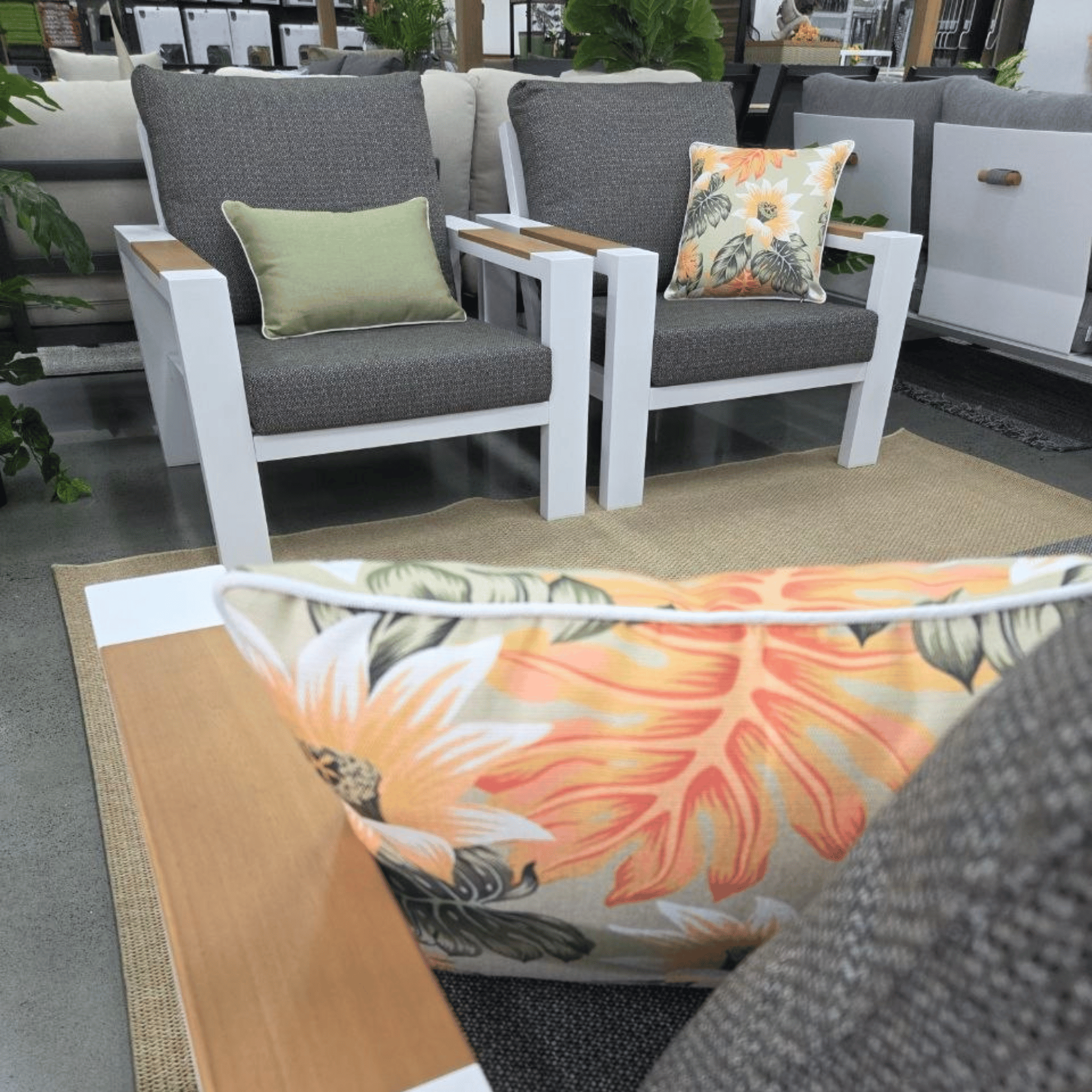 Whte outdoor patio armchairs with charcoal cushions and light wood accents, styled with decorative throw pillows in a showroom setting.
