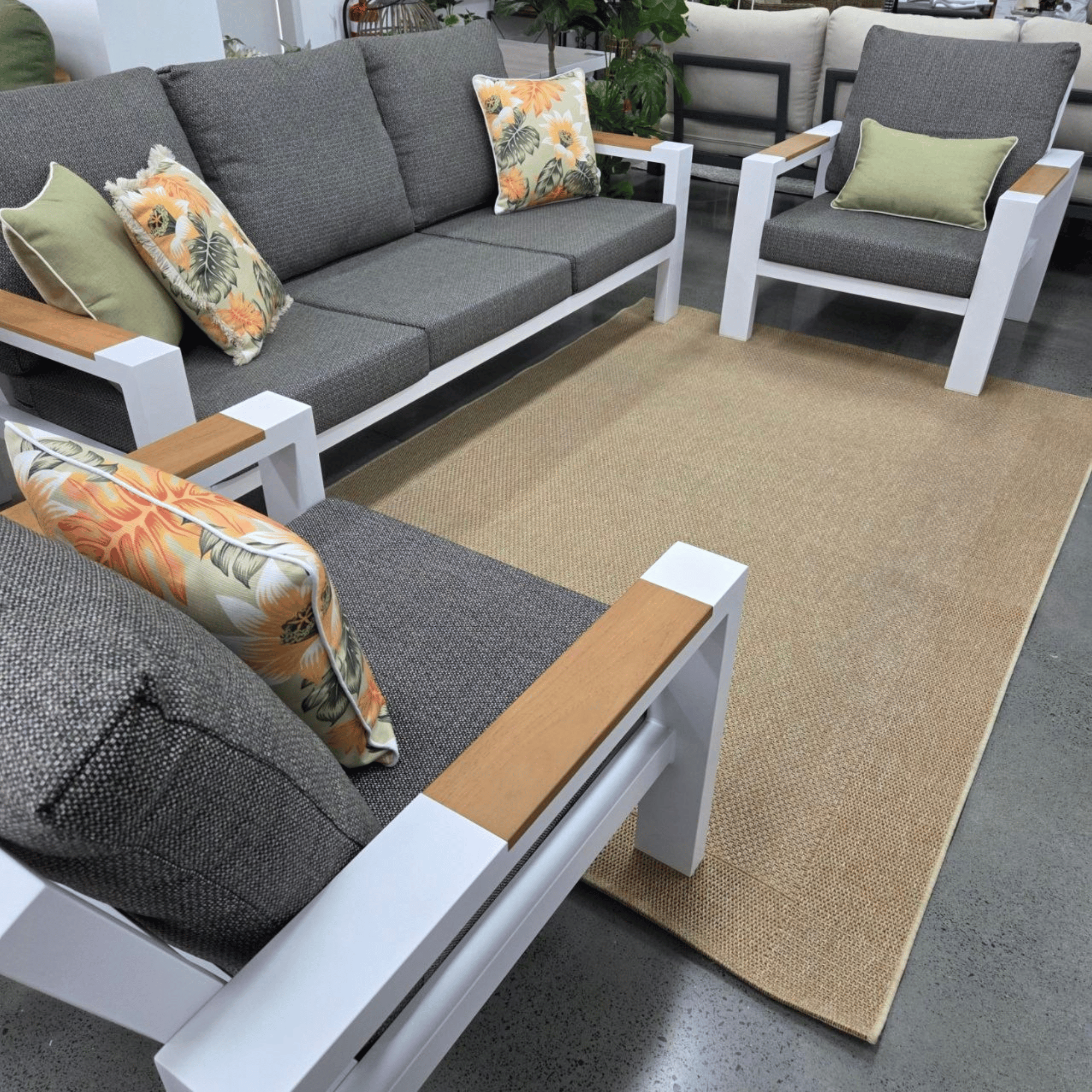 A modern Aspen 3pce outdoor lounge with grey textured cushions, white frames, and light wood armrests, arranged on a natural jute rug in a showroom.