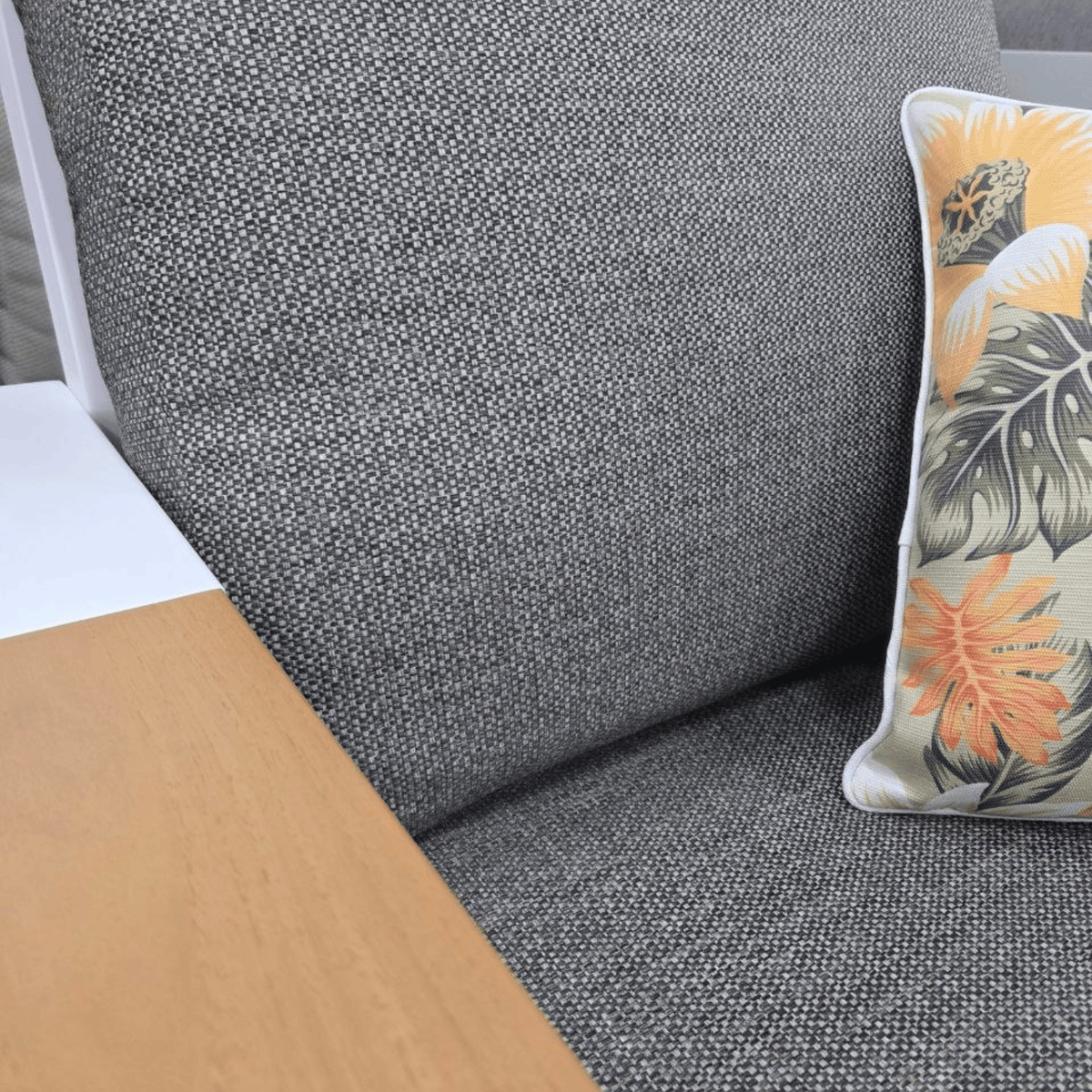 Close-up of an Aspen lounge with grey textured fabric sofa cushion with a decorative throw pillow featuring a tropical leaf and flower pattern in orange, beige, and green.