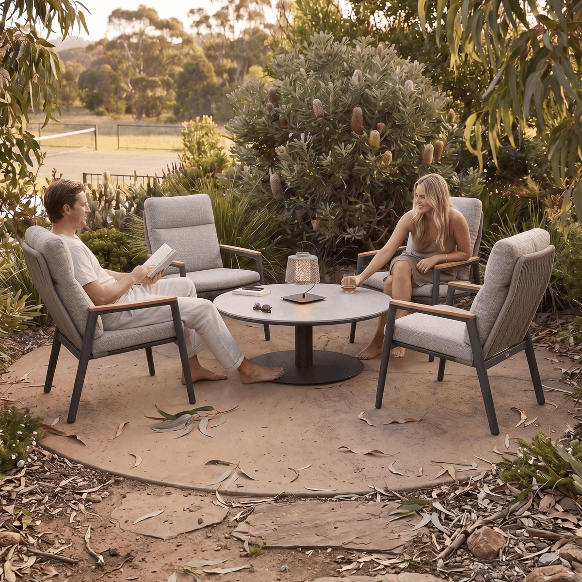 A couple sits in a backyard conversation area with four modern outdoor armchairs and a round coffee table. The setting is surrounded by a large bush, trees, and a dirt ground cover with scattered leaves.