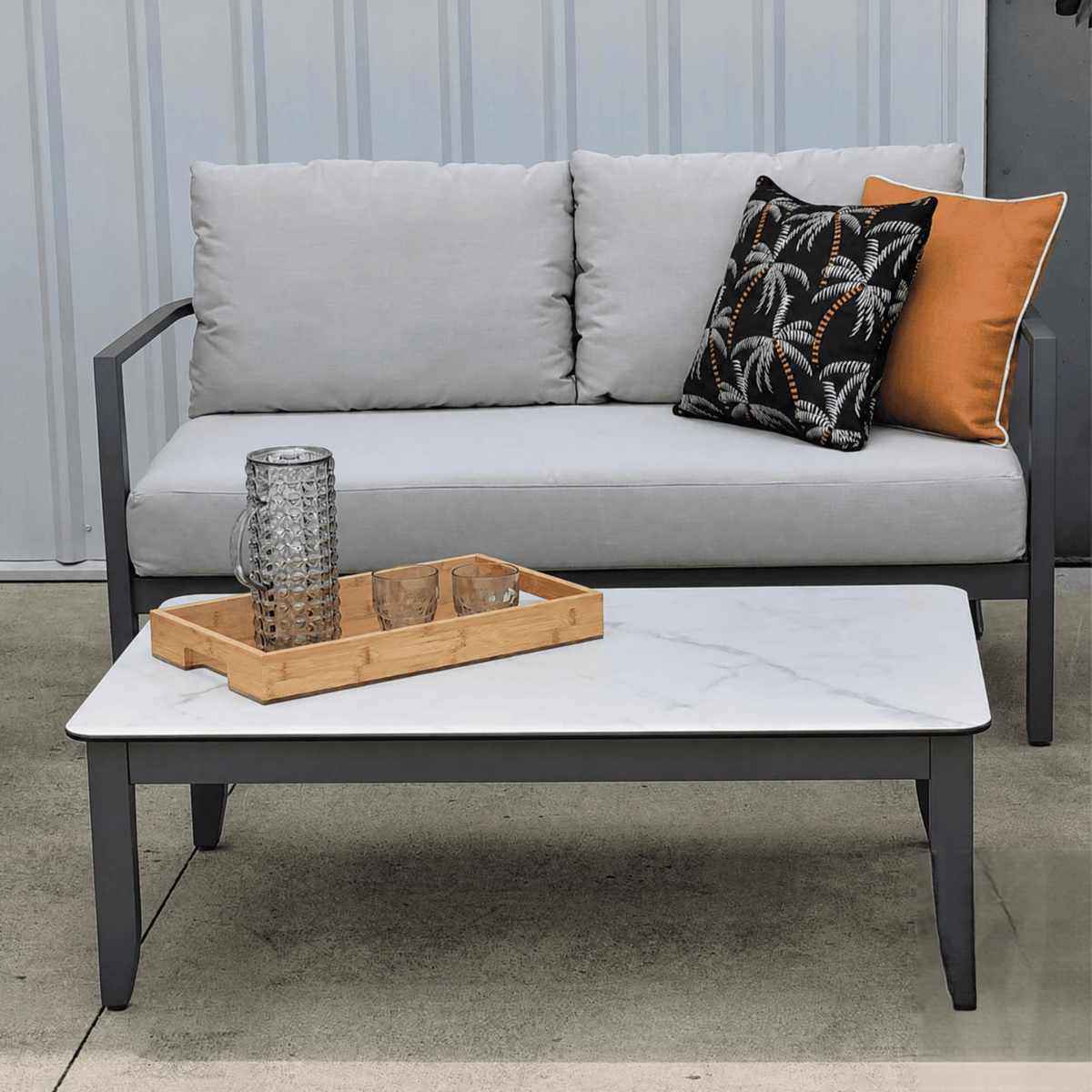 A modern outdoor patio set featuring a light gray two-seater sofa with dark gray metal frames, styled with a black palm-patterned throw pillow and an orange accent pillow. In front is a white marble-top coffee table with a dark gray frame, holding a bamboo tray with a textured glass tumbler.