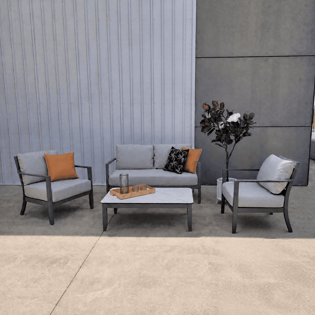 A 4-piece outdoor patio conversation set including a loveseat, two armchairs, and a coffee table, styled with gray cushions, orange accent pillows, and a potted plant against a modern gray paneled wall.