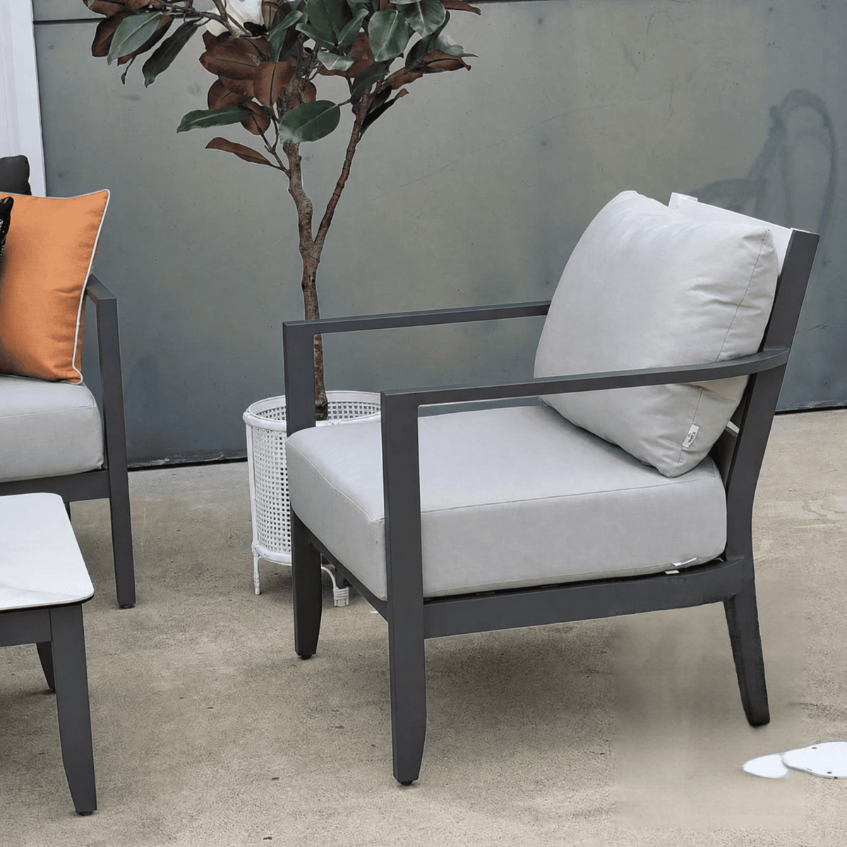 A modern outdoor armchair with a dark gray frame and light gray cushions, shown on a patio next to a matching side table and a potted plant.
