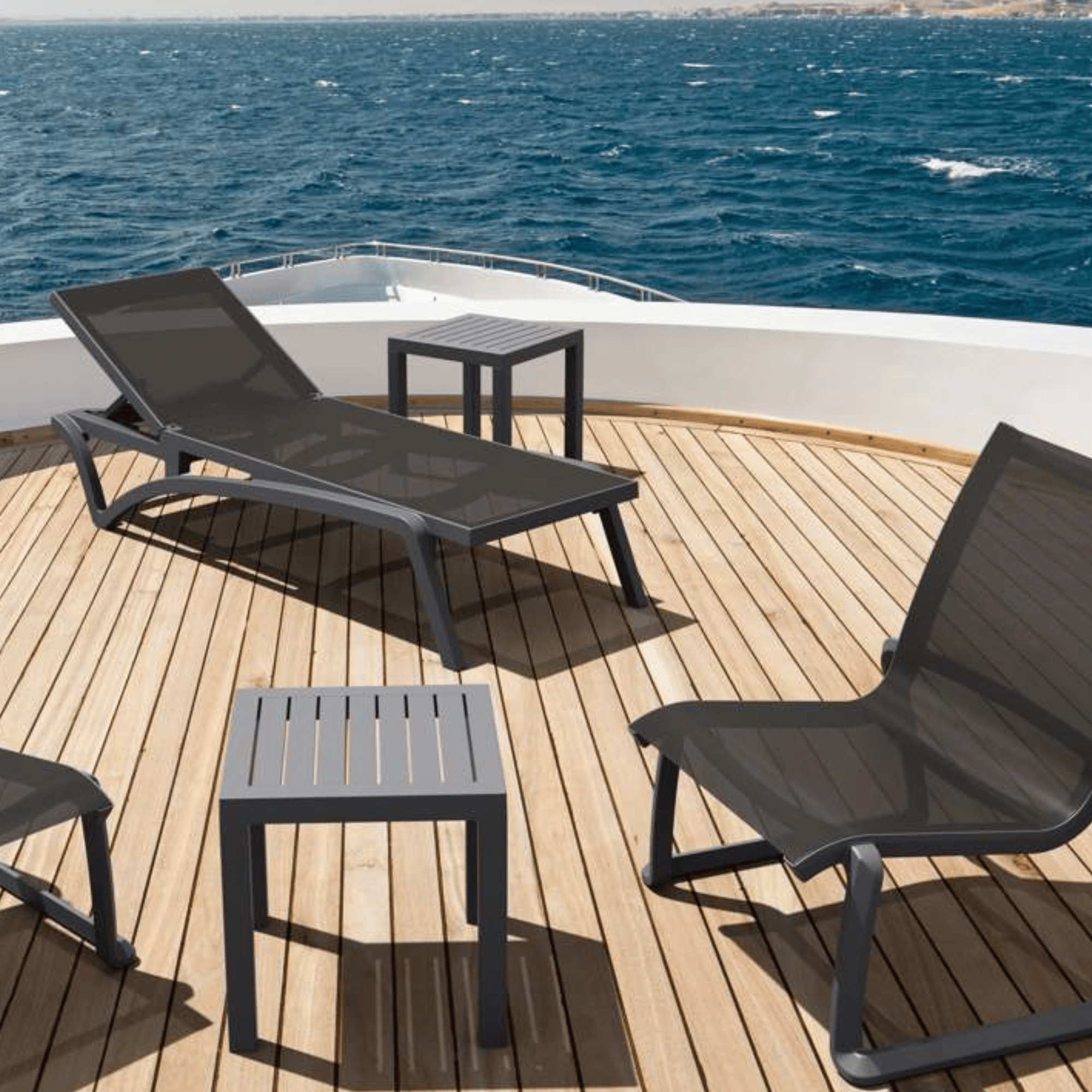 Outdoor yacht deck furniture set including Siesta Pacific Sunlounge - Anthracite - Commercial Grade, matching armchair, two small square slatted side tables, and a wooden deck with blue ocean water in the background.