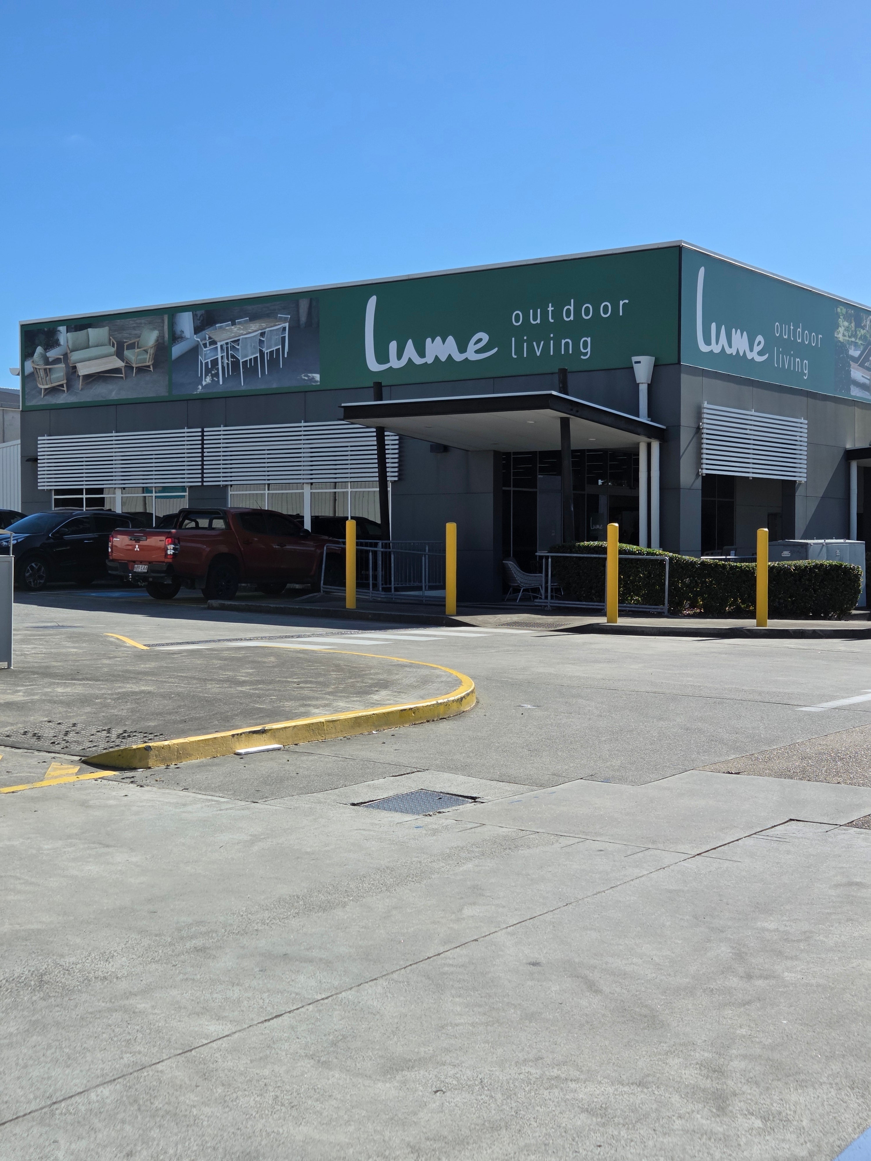 Lume Outdoor Living retail store exterior under a clear blue sky, featuring a green and gray building, parking area with vehicles, and yellow bollards.